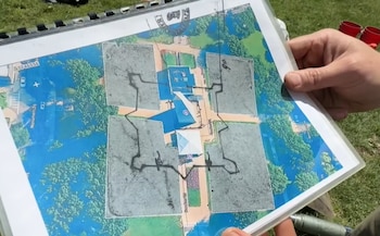 Manos sostienen un mapa impreso a color con zonas verdes de árboles, azules de agua o características, y áreas grises con contornos negros, posiblemente un sitio arqueológico