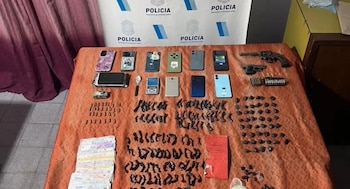 Objetos incautados por la policía: billetes, 10 celulares, dos armas de fuego y múltiples envoltorios negros con estupefacientes, sobre una mesa naranja y ante un cartel policial