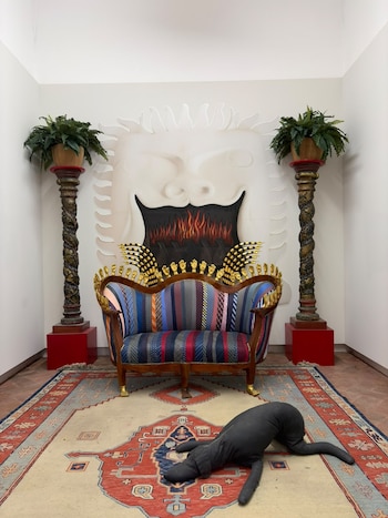 Un sofá a rayas con pequeñas manos doradas está sobre una alfombra junto a dos columnas con plantas y una figura negra en el suelo