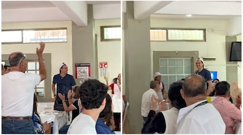 Un grupo de personas aplaude a un hombre sonriente con gorra blanca y camiseta oscura. Hay mesas, sillas y un código QR en la pared del salón.