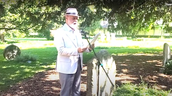 Un hombre de barba y gafas, vestido con sombrero blanco y chaqueta clara, habla por un micrófono al aire libre, sosteniendo papeles junto a una lápida de piedra