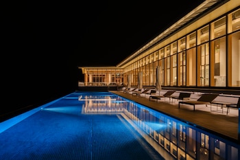 Piscina rectangular con luz azul junto a un edificio con ventanales de madera y vidrio iluminados por la noche con tumbonas en la terraza