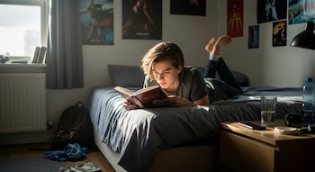 Un joven de cabello castaño claro yace boca abajo en su cama, leyendo un libro rojo. La luz del sol ilumina su rostro y la habitación.