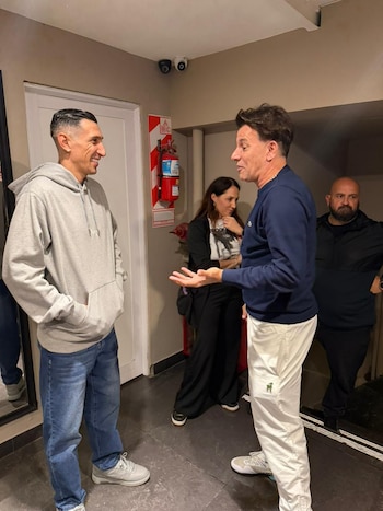 Ángel Di María con sudadera gris conversa de pie con Martín Bossi cerca de un extintor en un pasillo