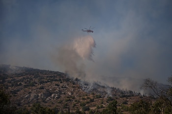 Un helicóptero rojo descarga un chorro de agua sobre una ladera rocosa y arbolada de la que emerge humo y fuego