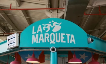 Primer plano de un letrero azul turquesa semicircular con el texto blanco 'LA MARQUETA' y un logo de dos figuras estilizadas. Cuelgan banderas de Puerto Rico y luces rosadas debajo, con cámaras de seguridad y un dispositivo blanco de control de insectos a los lados