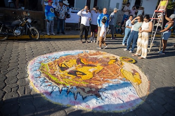 Un hombre con gorra azul crea una alfombra de aserrín colorida en el suelo, con diseños de un sol radiante y dos estructuras arquitectónicas coloniales, en una calle empedrada