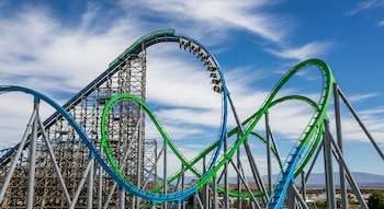 Vista amplia de la montaña rusa Twisted Colossus con sus vías entrelazadas azules y verdes, estructura de soporte de madera y acero bajo un cielo azul con nubes dispersas.
