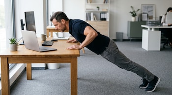 Un hombre con las manos sobre un escritorio de madera realiza una flexión inclinada junto a una computadora portátil y una pantalla.