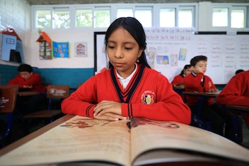 Niña con suéter rojo lee un libro abierto sobre un pupitre de madera en un aula. Al fondo se ven otros estudiantes, paredes blancas y ventanas