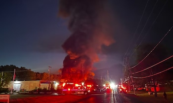 Incendio nocturno en edificio industrial con gran columna de humo y llamas. Vehículos de emergencia y personal en la calle iluminada por luces rojas