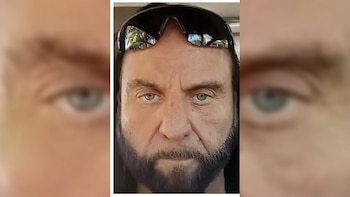 Primer plano de un hombre caucásico de mediana edad con barba oscura y pelo largo, llevando gafas de sol negras en la frente y mirando al frente