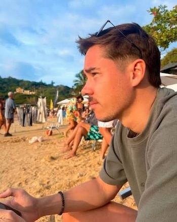 Hombre joven de perfil con gafas de sol sobre la cabeza, sentado en la arena de una playa. Al fondo, otras personas, sombrillas y puestos de venta