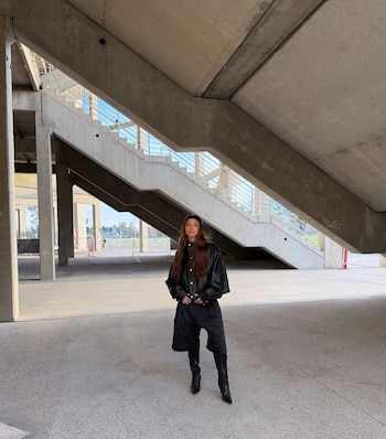 Ángela Torres de pie bajo una escalera de hormigón. Viste chaqueta de cuero negra, pantalón bermuda negro y botas altas. Hay barandas metálicas