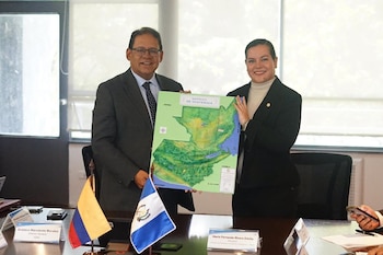 Un hombre de traje y una mujer con blazer sostienen un mapa de la República de Guatemala. En una mesa hay banderas de Colombia y El Salvador y credenciales
