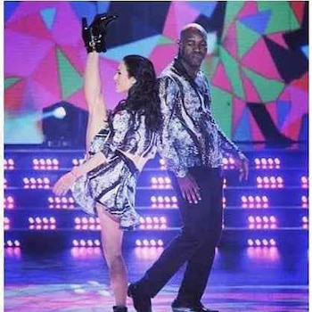 Una mujer con vestido estampado levanta una pierna verticalmente junto a un hombre con camisa a juego en un escenario iluminado