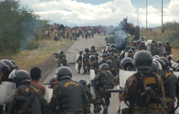 Agentes de la Policía Nacional con cascos y escudos avanzan por una carretera rural con humo. Al fondo, una multitud de manifestantes y un vehículo blindado