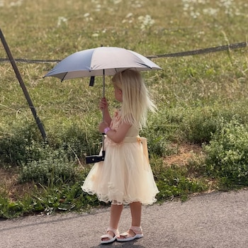 Niña rubia de perfil con vestido claro, sandalias y pulsera rosa, sosteniendo un paraguas gris. Está en un camino junto a un campo con vegetación