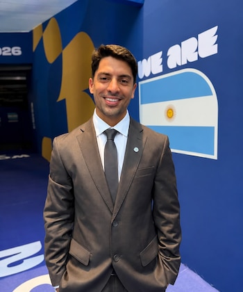 Hombre sonriendo, traje gris, camisa blanca, corbata gris, pin en solapa, fondo azul con formas amarillas, bandera de Argentina, texto WE ARE, numero 2026