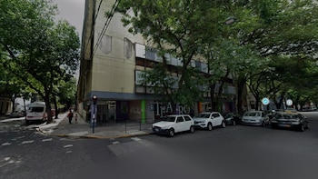Esquina de calle con edificio de fachada clara y árboles. Varios automóviles estacionados, un semáforo rojo. Dos personas y una camioneta blanca en la acera