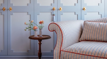 Sofá tapizado con estampado de rayas y puntos, mesa auxiliar de madera con dos floreros, ranúnculos rosados, y pared panelada con tiradores dorados y un borlón.