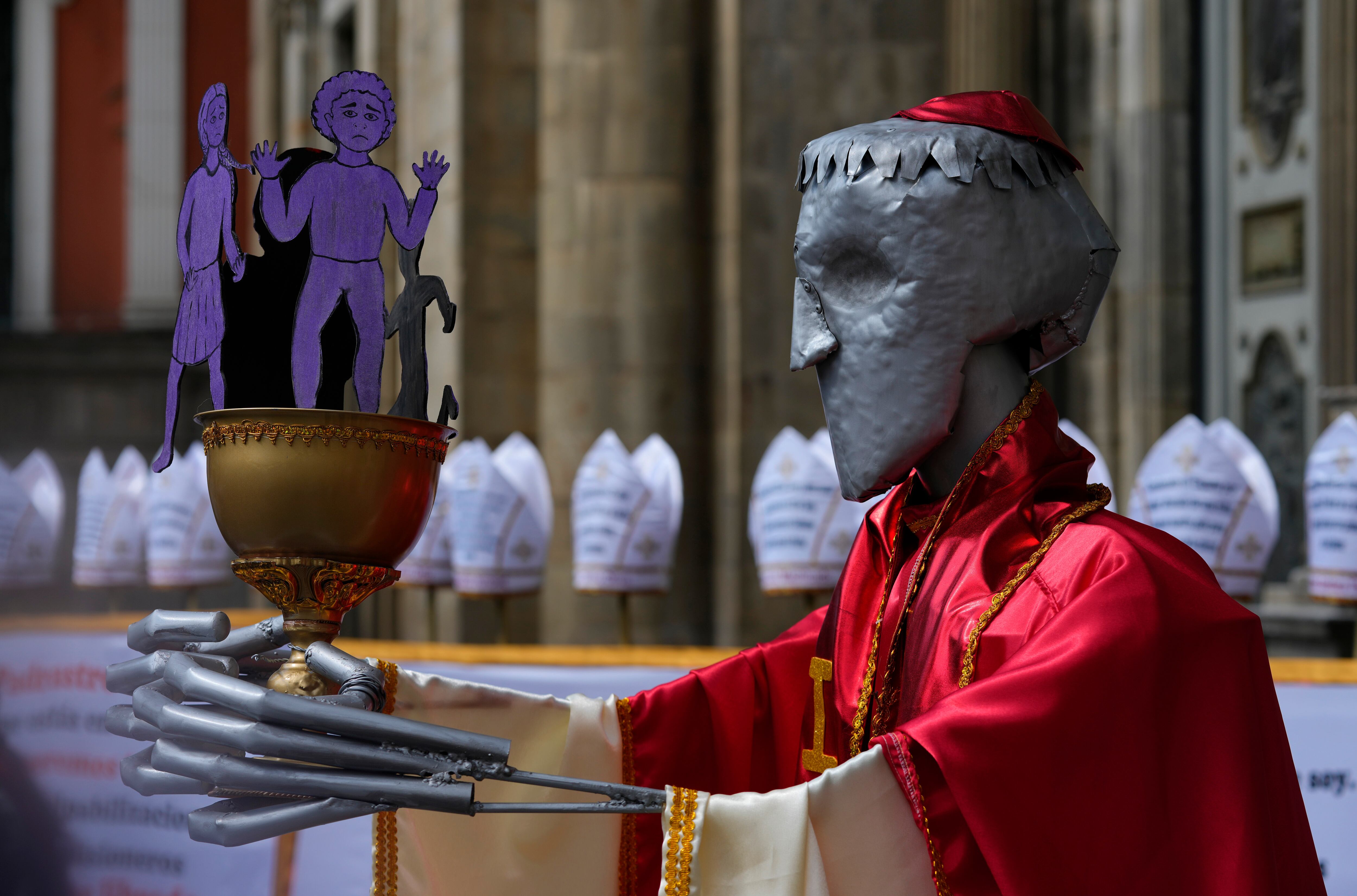 Una figura que representa a un cura católico con cáliz lleno con recortes con forma de niños colocada junto a varias mitras con mensajes contra los abusos sexuales a menores, en una protesta organizada por el grupo feminista Mujeres Creando, en reclamo de condenas para los sacerdotes acusados de pederastia, en las afueras de la Catedral Metropolitana en La Paz, Bolivia, el jueves 16 de mayo de 2024. (AP Foto/Juan Karita)