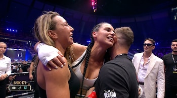 Dos mujeres con vestimenta deportiva y un hombre con camiseta negra se abrazan en un escenario iluminado, con espectadores y un contador de audiencia en pantalla.