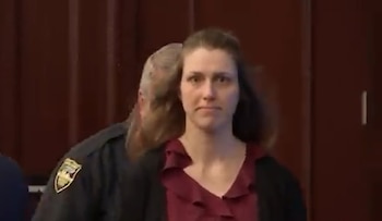 Mujer de cabello castaño y blusa roja con volantes mirando al frente. Detrás, un oficial de seguridad uniformado con un escudo en el hombro