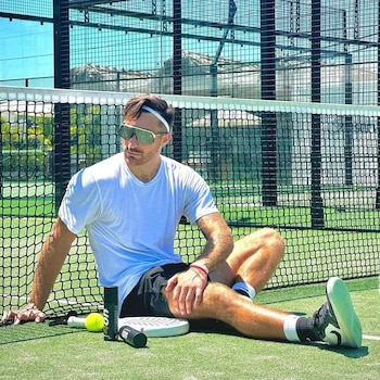 Hombre sentado en el suelo de una cancha de pádel verde, con camiseta blanca, pantalones cortos negros, gafas de sol y cinta de pelo, junto a una red, raqueta, pelota y dos envases