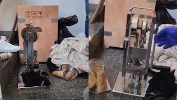 Una prensa metálica manual en el suelo junto a una caja de madera y una mano con guante azul en una imagen de dos tomas