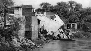 Fotografía en blanco y negro que muestra edificios parcialmente colapsados, escombros en la orilla del río y árboles con vegetación densa