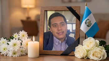 Un retrato enmarcado de un joven con una cinta negra, una vela encendida, flores blancas y una bandera de Guatemala sobre una mesa de madera.
