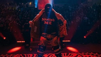 Un individuo enmascarado con gorro negro y gafas de sol abre su abrigo sobre una camiseta que dice "Abolish ICE" en un escenario iluminado con luces rojas y azules, frente a una multitud oscura
