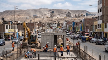Obreros con cascos y chalecos trabajan en una obra con una excavadora, entre edificios comerciales, vehículos y cerros con maquinaria.
