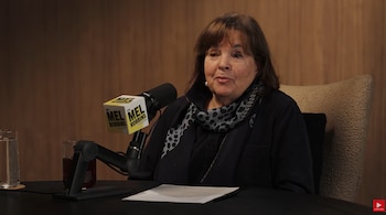 Una mujer sentada ante un micrófono con soporte, con papeles y dos vasos sobre una mesa oscura frente a un panel de madera