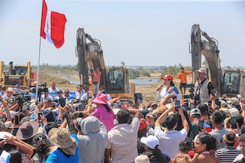 Una multitud de personas con teléfonos móviles asiste a un evento al aire libre con maquinaria pesada, una bandera peruana y un río al fondo