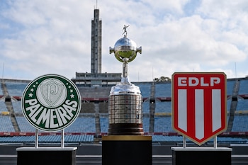 El trofeo de la Copa Libertadores entre el escudo verde de Palmeiras y el escudo rojo de Estudiantes de La Plata frente a las gradas de un estadio.