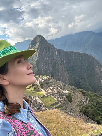 Mujer con sombrero verde y camisa estampada mirando hacia arriba, con las ruinas de Machu Picchu y montañas al fondo