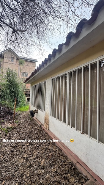 Exterior de una casa con fachada blanca y techo de tejas rojas. Ventanas enrejadas y suelo cubierto de hojas secas. Árboles sin hojas y otra casa al fondo