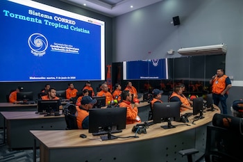 Sala de control con personas en chalecos naranjas sentadas frente a monitores, escuchando a un orador. Una pantalla grande muestra "Tormenta Tropical Cristina"