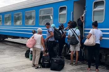 Varios pasajeros con mochilas y bolsos esperan y abordan la puerta de un vagón de tren azul en un andén.
