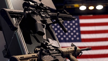 Dos rifles de asalto, uno negro y otro color arena, se muestran en un estante perforado, con la bandera de Estados Unidos en el fondo desenfocado