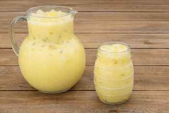Una jarra de cristal transparente llena de una bebida amarilla con trozos de fruta y cubitos de hielo, junto a un vaso también de cristal con la misma bebida, ambos sobre una superficie de madera.