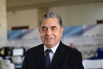 Retrato de un hombre de edad avanzada con cabello canoso, vistiendo traje oscuro, camisa blanca y corbata azul estampada, sonriendo a la cámara