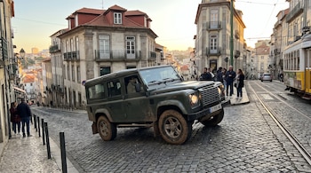 Vehículo todoterreno verde oscuro con barro sobre calle adoquinada, edificios históricos, tranvía amarillo, personas, postes y cielo al atardecer.