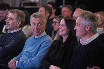 Mauricio Macri con suéter azul claro sonríe sentado junto a una mujer de cabello oscuro y un hombre de cabello gris, también sentados en una audiencia