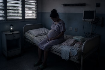Una mujer embarazada se sienta en una cama de hospital junto a ropa de bebé, pañales, un monitor apagado y un farol encendido.