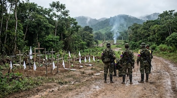 Soldados colombianos en un camino de tierra observan un cementerio improvisado de cruces de madera y banderas blancas en la selva, con humo de fondo.