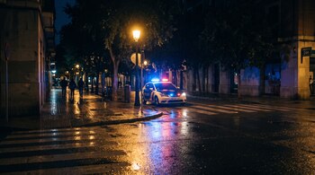 Un automóvil policial con luces de emergencia encendidas junto a un paso de peatones en una calle mojada e iluminada por farolas.