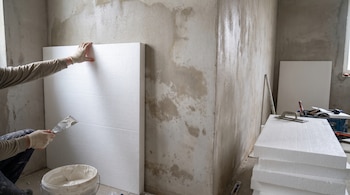Un trabajador con guantes sujeta un panel de poliestireno expandido blanco en una pared, con una espátula y una cubeta de adhesivo cerca. Hay paneles apilados.